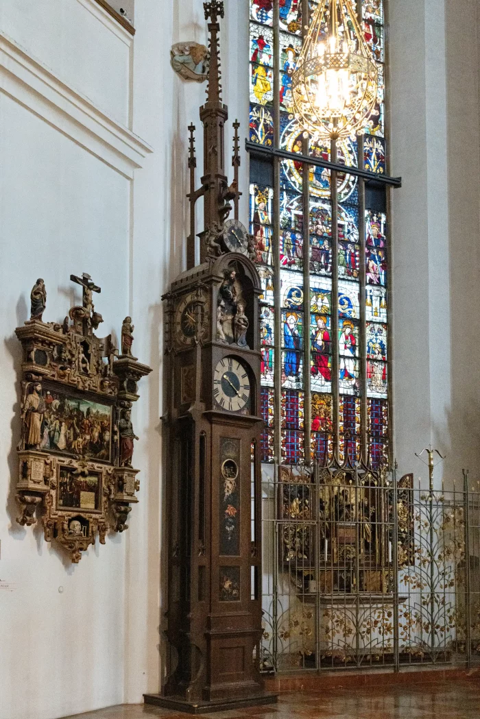 Hohe Standuhr mit geschnitztem Holzgehäuse und Zifferblatt, daneben geschnitztes Wandrelief mit Kreuz, im Hintergrund buntes Kirchenfenster der Frauenkirche München.