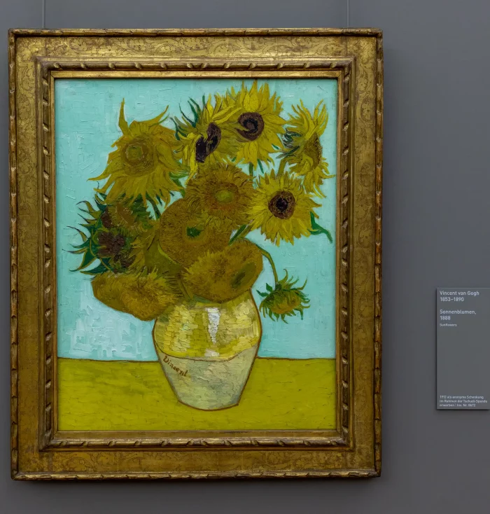 Gemälde von Vincent van Gogh mit Sonnenblumen in einer hellen Vase vor blauem Hintergrund, gerahmt in einem goldfarbenen Rahmen.