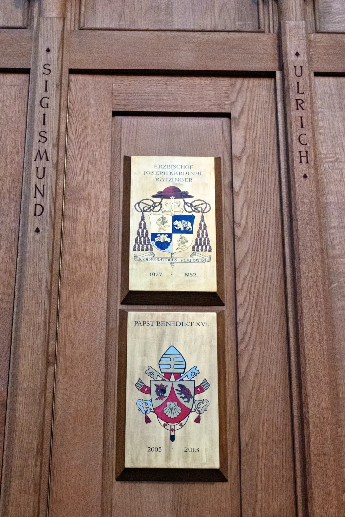 Zwei Wappentafeln an Holztür, oben mit Text 'Erzbischof Josef Kardinal Ratzinger 1977 1982', unten 'Papst Benedikt XVI. 2005 2013', links und rechts die Namen 'Sigismund' und 'Ulrich'.