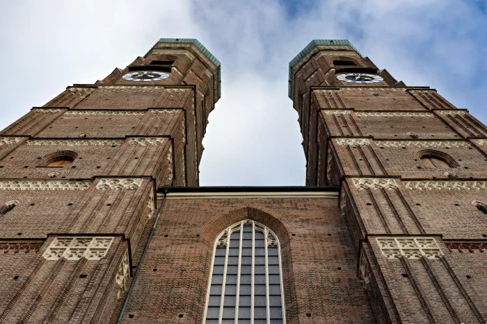 Blick von unten auf die zwei Backsteintürme der Frauenkirche in München mit Uhr und spitzbogigen Fenstern vor bewölktem Himmel.