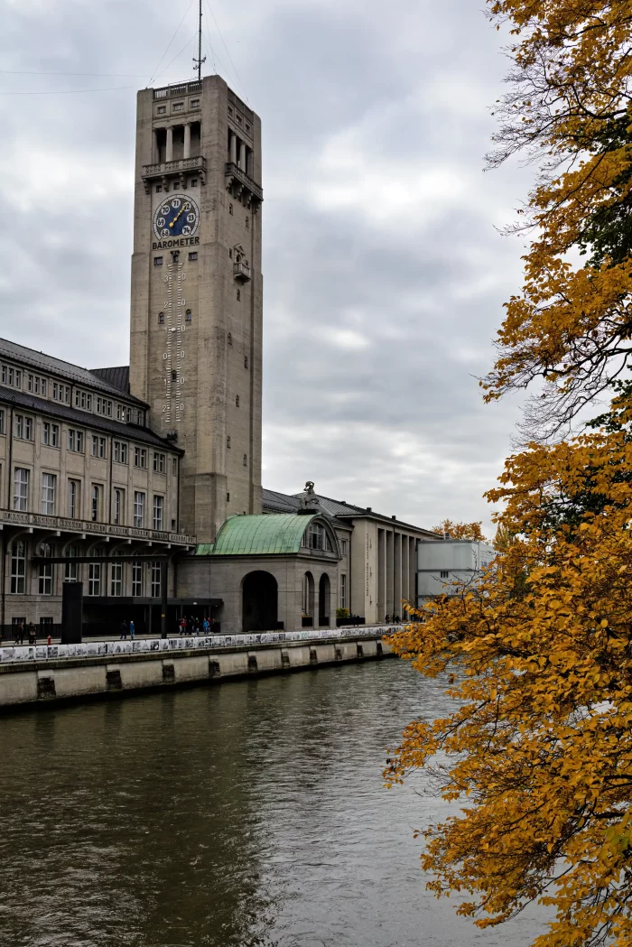 Der markante Uhrenturm des Deutschen Museums in München neben einem Fluss, rechts Herbstlaub an einem Baum.