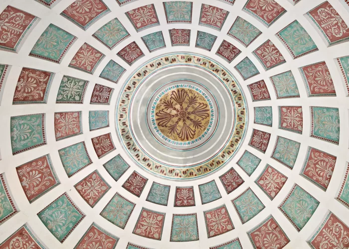 Blick nach oben auf die bemalte Kuppel im Englischen Garten München mit konzentrischen Ringen aus rot- und türkisfarbenen Quadraten und einem goldfarbenen Mittelkreis mit floralem Muster.