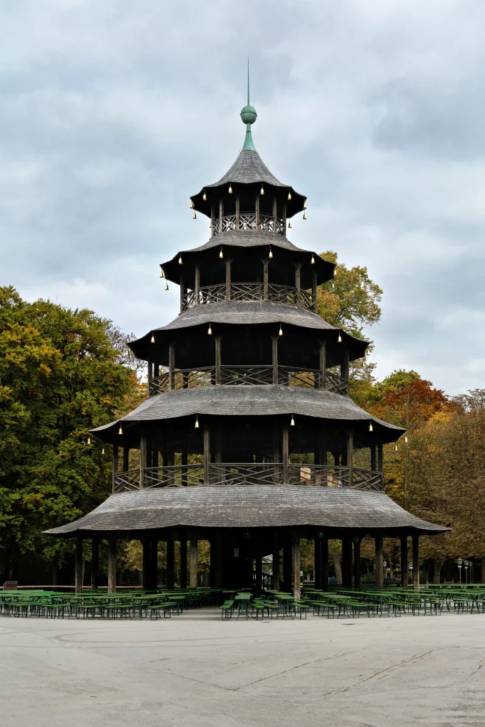 Fünfstöckiger, offener Holzturm mit spitzem Dach und grünem Kugelaufsatz im Englischen Garten in München, umgeben von Bäumen.