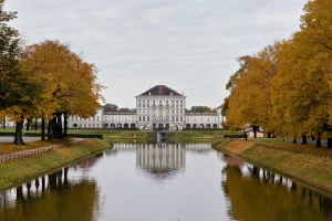 Schloss Nymphenburg in München, frontal mit Wassergraben und herbstlich gefärbten Bäumen auf beiden Seiten.