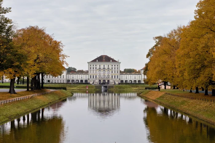 Schloss Nymphenburg in München, frontal mit Wassergraben und herbstlich gefärbten Bäumen auf beiden Seiten.