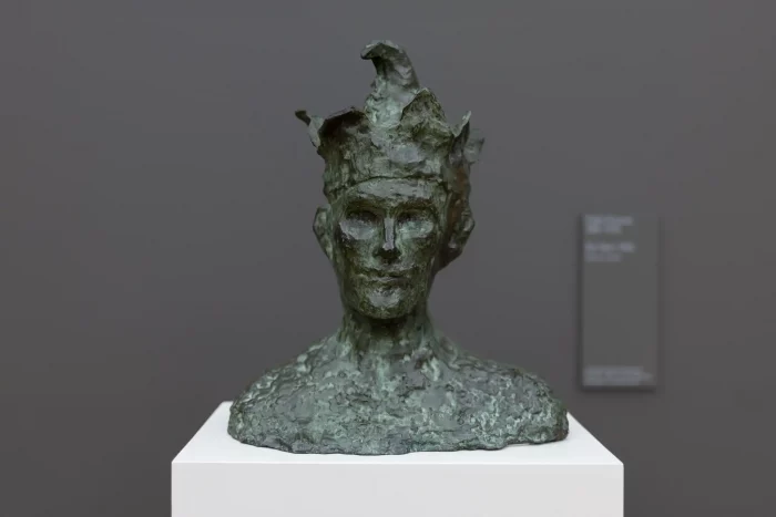 Bronzebüste mit unregelmäßiger, rauer Oberfläche und spitzen Formen auf einem weißen Sockel vor grauer Wand.