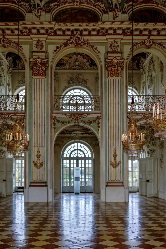 Großer Saal im Schloss Nymphenburg mit hohen verzierten Säulen, großen Rundbogenfenstern und Kronleuchtern, Parkettboden mit Schachbrettmuster.