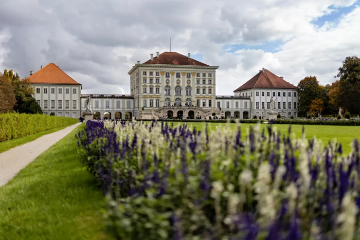 Schloss Nymphenburg in München mit barockem Mittelbau, zwei Seitenflügeln und Blumenbeeten im Vordergrund unter bewölktem Himmel.