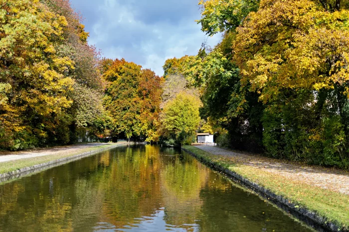 Herbstliche Bäume mit buntem Laub entlang eines ruhigen Kanals im Schlosspark Nymphenburg in München.