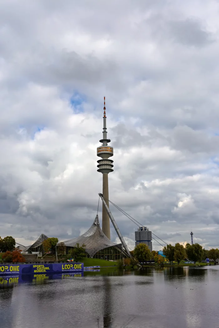Olympiaturm im Olympiapark München mit markanter Antenne, davor Zeltdachkonstruktionen und Wasserfläche, bewölkter Himmel.