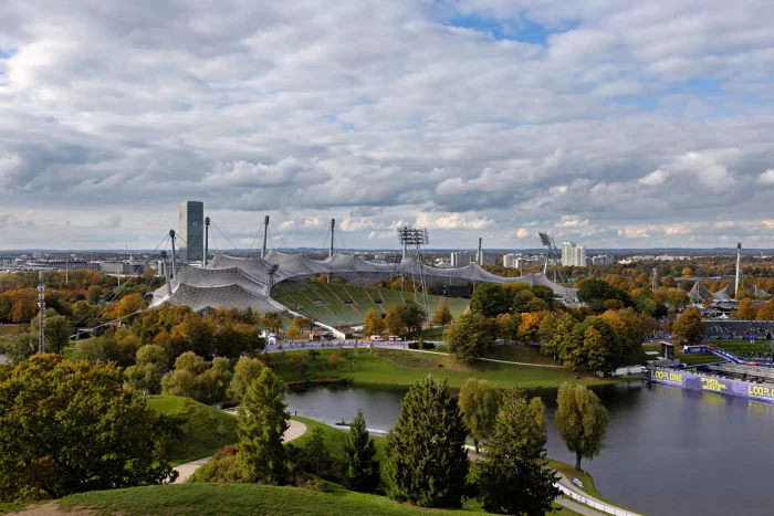 Aufnahme des Olympiaparks München mit dem markanten Zeltdach des Olympiastadions, umgeben von herbstlich gefärbten Bäumen und einem See im Vordergrund.