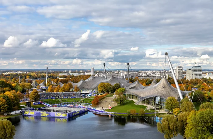 Blick auf den Olympiapark München mit dem Zeltdach des Olympiastadions, herbstlich gefärbten Bäumen und einem See im Vordergrund.