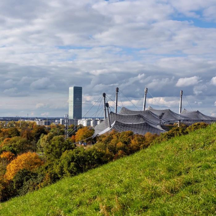Blick auf das Zeltdach des Olympiaparks München mit herbstlich gefärbten Bäumen und dem markanten Bürohochhaus im Hintergrund unter bewölktem Himmel.