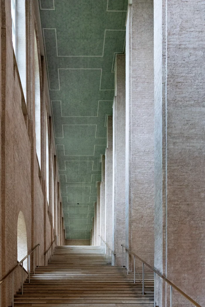 Blick in einen hohen, schmalen Treppenraum mit weißen Wänden und grünem, geometrisch gemustertem Deckenbereich.