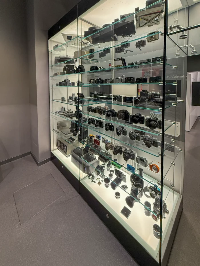 Vitrine mit mehreren Regalböden, darauf zahlreiche verschiedene Kameras und Objektive in einem Ausstellungsraum.