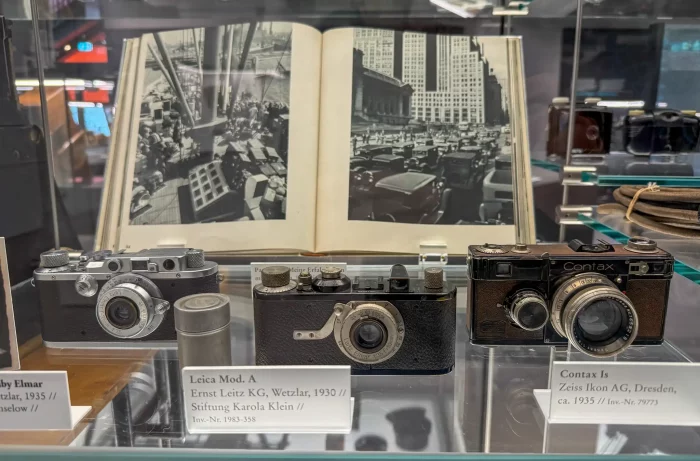 Drei historische Kameras von Leica, Contax und Zeiss Ikon auf Glasvitrine mit offenem Buch im Hintergrund, das schwarz-weiße Stadt- und Straßenszenen zeigt.