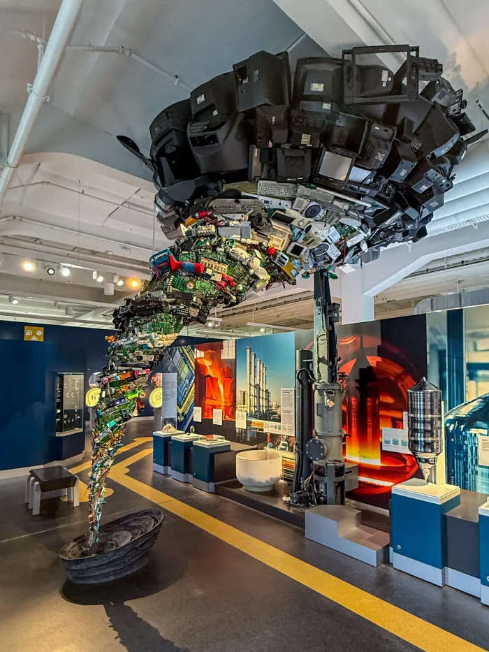 Skulptur aus alten Fernsehern, Radios und Elektronikgeräten, die sich spiralförmig von einem runden Sockel bis zur Decke windet.