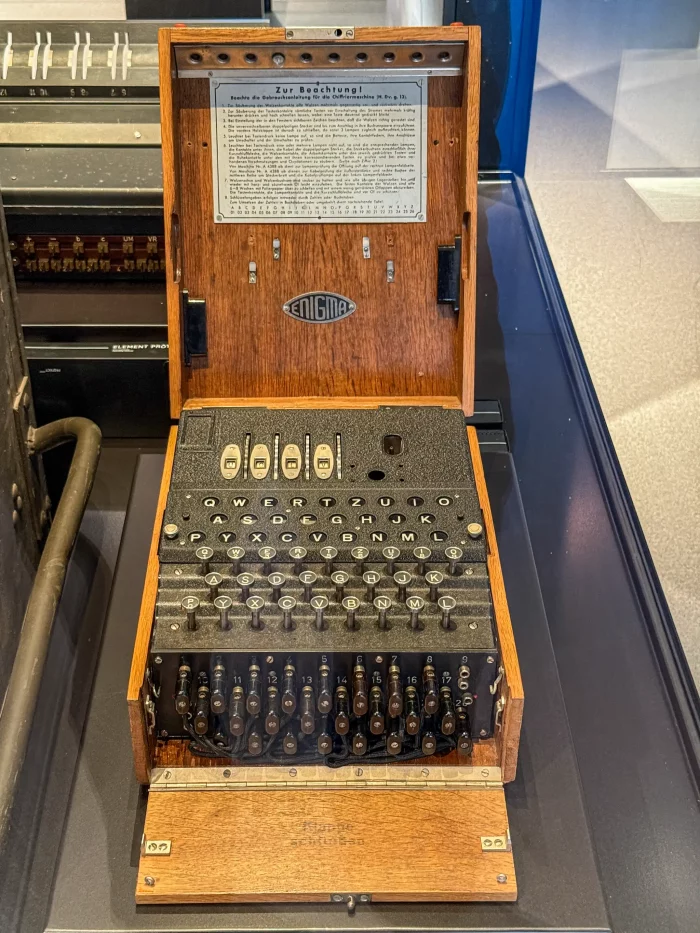 Offene Enigma-Chiffriermaschine aus Holz mit Tastatur, Schaltern und Metallhebeln, darunter ein Hinweisblatt mit deutscher Beschriftung.