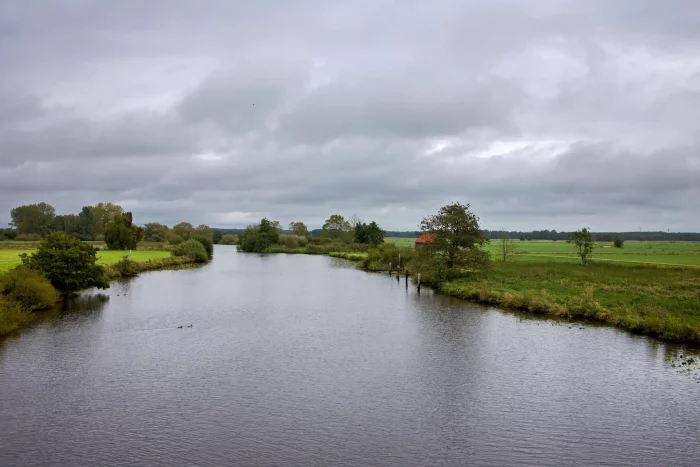 Eine ruhige Landschaft mit einem gewundenen Fluss, umgeben von grasbewachsenen Ufern und Bäumen. Der Himmel ist teilweise bewölkt, mit einem Hauch von Blau, der durchschaut. Ein kleines rotes Gebäude ist in der Ferne nahe dem Wasserrand zu sehen.