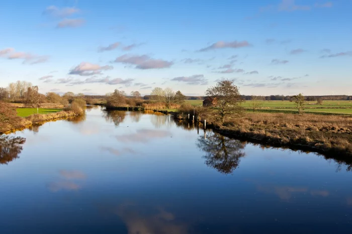 Eine ruhige Landschaft mit einem gewundenen Fluss, umgeben von grasbewachsenen Ufern und Bäumen. Der Himmel ist teilweise bewölkt, mit einem Hauch von Blau, der durchschaut. Ein kleines rotes Gebäude ist in der Ferne nahe dem Wasserrand zu sehen.