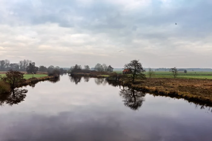 Eine ruhige Landschaft mit einem gewundenen Fluss, umgeben von grasbewachsenen Ufern und Bäumen. Der Himmel ist teilweise bewölkt, mit einem Hauch von Blau, der durchschaut. Ein kleines rotes Gebäude ist in der Ferne nahe dem Wasserrand zu sehen.