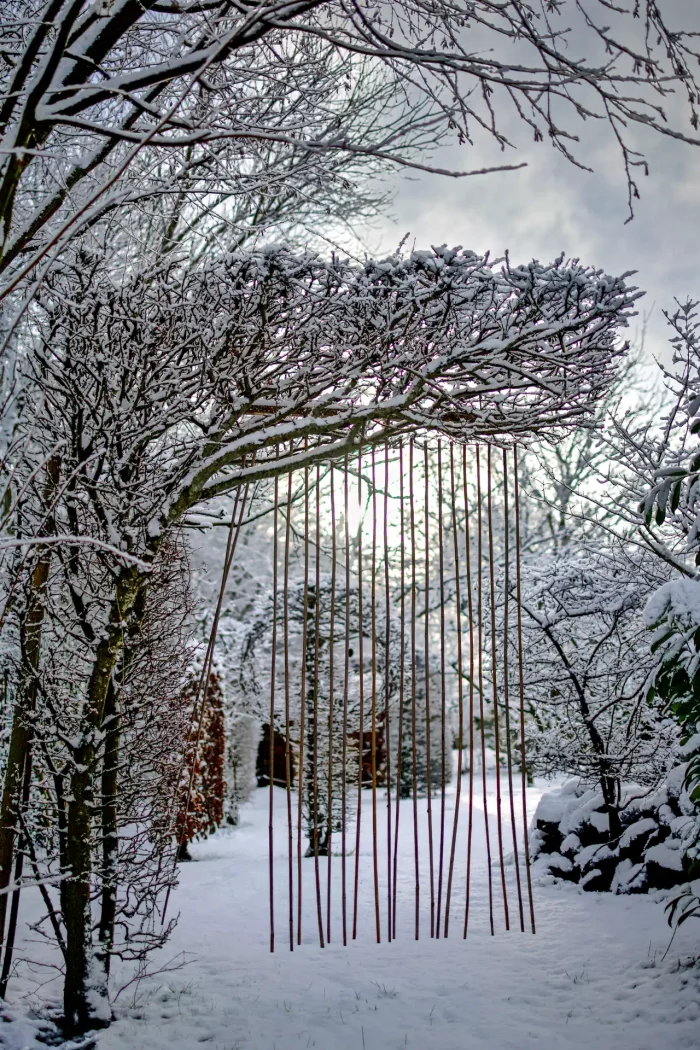 Schneebedeckter Gartenweg mit kahlen Bäumen und einer Installation aus vertikal hängenden Stäben unter einem schneebedeckten Astbogen