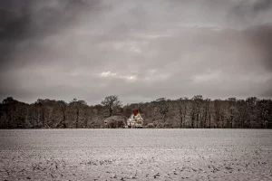 Verschneites Feld mit einem einzelnen Haus und kahlen Bäumen unter bewölktem Himmel