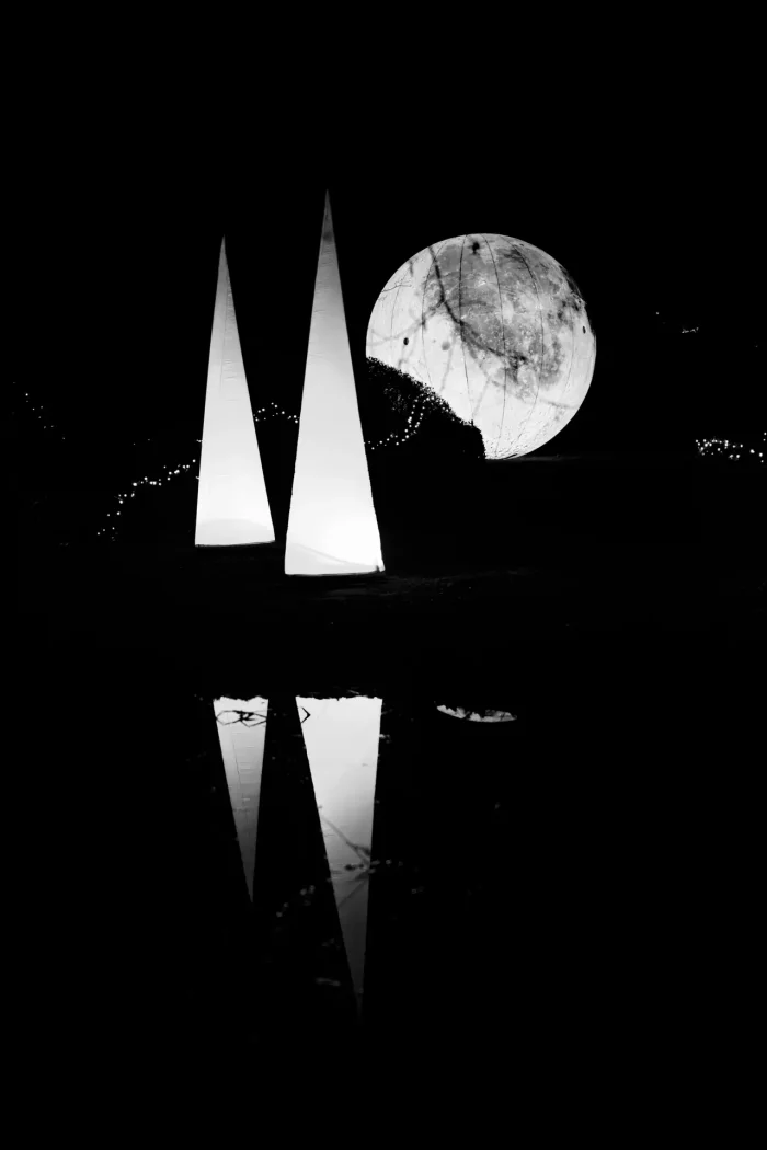 Zwei beleuchtete Pyramiden und eine große beleuchtete Kugel mit Baumzweigen im Hintergrund und deren Spiegelung im Wasser