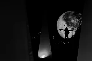 Silhouette einer Person mit ausgebreiteten Armen vor einem großen, beleuchteten Mond in der Nacht