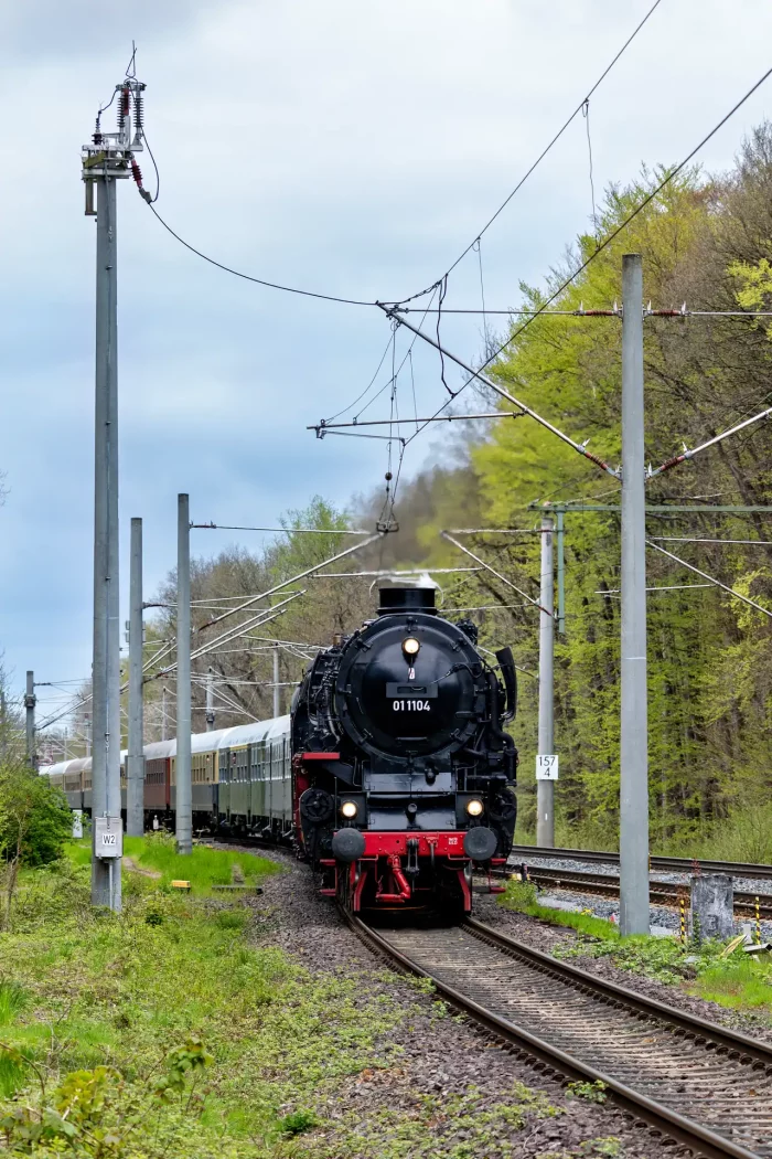 Historische Dampflok 011104 zieht einen Zug auf einer elektrifizierten Strecke mit Oberleitungen vor bewaldetem Hintergrund bei Lübberstedt
