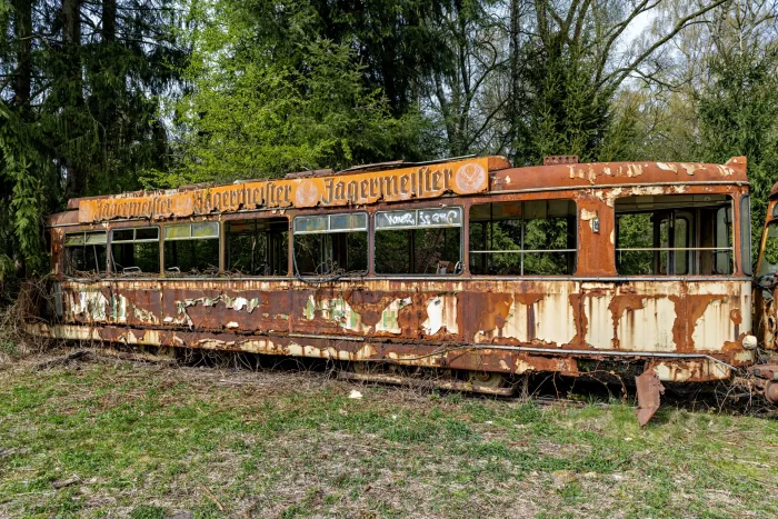 Verfallene Straßenbahn mit abblätterndem Lack und verblasster Werbung für Jägermeister steht auf einer Wiese vor Nadelbäumen