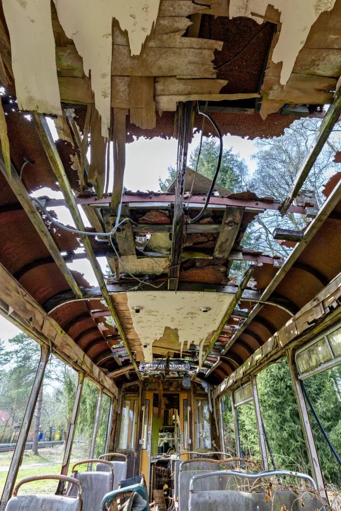 Innenraum einer verfallenen Straßenbahn mit teilweise fehlender und herabfallender Deckenverkleidung, sichtbaren Kabeln und rostigen Metallstreben, mehrere Sitzreihen und Fenster mit Blick auf Bäume im Freien