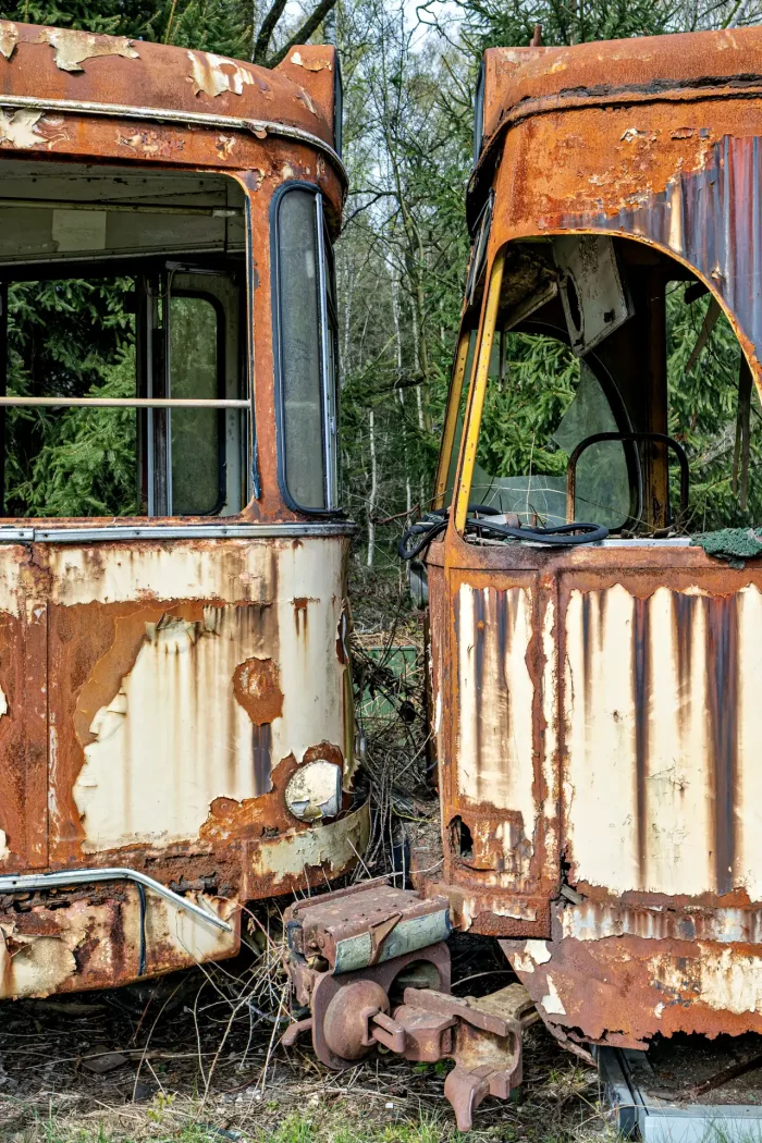 Zwei verrostete, verfallene Straßenbahnwagen stehen dicht nebeneinander in einem bewaldeten Lostplace