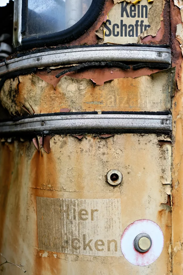 Nahaufnahme einer verfallenen Straßenbahn mit abgeblättertem Lack, rostigen Metallstreifen, verblasstem Schild mit der Aufschrift 'Hier drücken' und einem kleinen runden Knopf sowie einem weiteren Schild mit der Aufschrift 'Kein Schaffner'.
