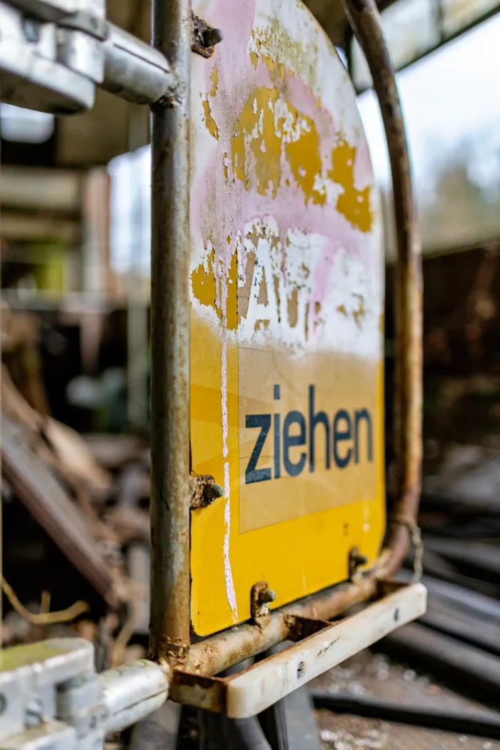 Nahaufnahme eines verrosteten Metallschilds an einer verfallenen Straßenbahn mit verblasstem Aufdruck und dem gut lesbaren Wort 'ziehen' auf gelbem Hintergrund