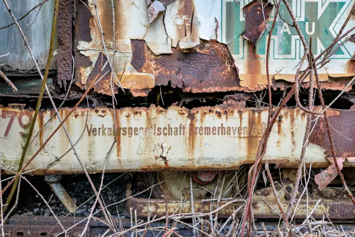 Nahaufnahme eines stark verrosteten und verfallenen Teils einer alten Straßenbahn mit verblasster Aufschrift 'Verkehrsgesellschaft Bremerhaven AG' und überwucherten Ästen