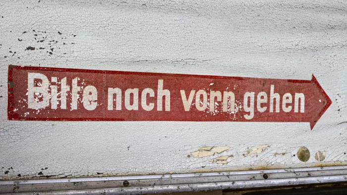 Rostige, abgeblätterte Wand mit rotem Pfeil und weißer Schrift 'Bitte nach vorn gehen' in einer verfallenen Straßenbahn.