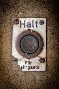 Verrosteter, rechteckiger Metallknopf mit der Aufschrift 'Halt Für Fahrgäste' auf einer rissigen, braunen Oberfläche befestigt