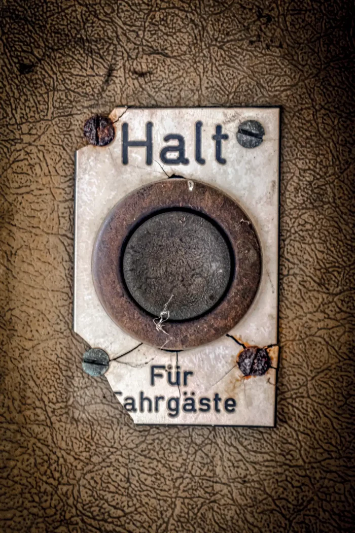 Verrosteter, rechteckiger Metallknopf mit der Aufschrift 'Halt Für Fahrgäste' auf einer rissigen, braunen Oberfläche befestigt