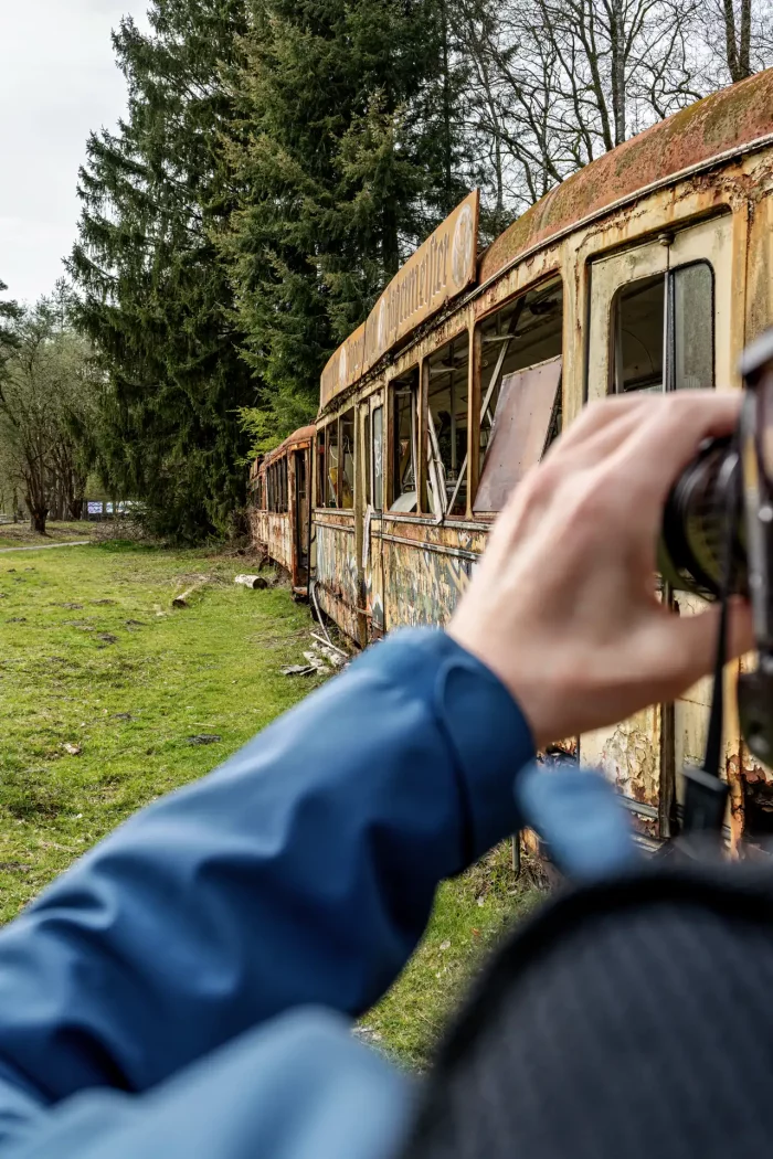 Verfallene Straßenbahn mit zerbrochenen Fenstern steht auf Grasfläche neben Nadelbäumen, im Vordergrund Hand mit Kamera in blauer Jacke