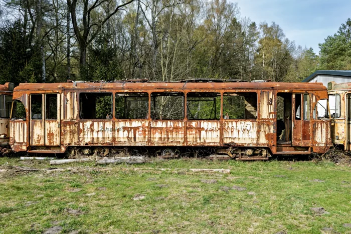 Verfallene Straßenbahn mit stark verrosteter Außenhülle und fehlenden Fensterscheiben steht auf Grasfläche vor Wald