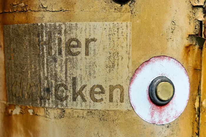 Verrostete Metallfläche mit verblasstem Schild 'Hier drücken' und rundem Knopf mit rotem Rand