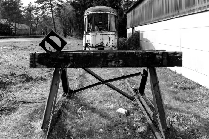 Verfallene Straßenbahn hinter einem alten Gleisabschluss mit schrägem Verkehrsschild neben einer Wand und Bäumen im Hintergrund