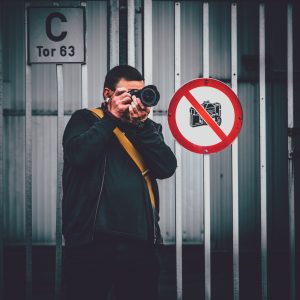 Person mit schwarzer Kleidung und gelbem Schulterriemen fotografiert vor Metalltor mit Schild 'C Tor 63' und Verbotsschild für Kameras