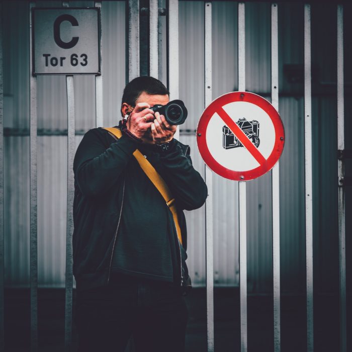 Person mit schwarzer Kleidung und gelbem Schulterriemen fotografiert vor Metalltor mit Schild 'C Tor 63' und Verbotsschild für Kameras