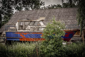 Ein blau-orange gestrichenes Boot steht auf dem Land vor einem Haus mit einem Ziegeldach und ist von Büschen umgeben