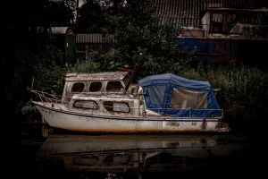 Altes, weißes Boot mit blauem Verdeck liegt am Ufer vor dunklem, bewachsenem Hintergrund