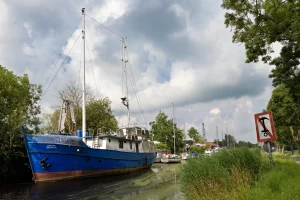 Blaues Segelschiff am Ufer eines Kanals mit mehreren weiteren Booten und einem Schild mit Anker-Symbol am rechten Bildrand