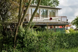Zweistöckiges Haus mit großem Balkon und einem langen weißen Kajak als Dekoration, umgeben von dichtem Grün und Bäumen