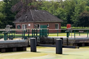 Backsteingebäude mit Satteldach hinter einer Schleuse mit grünen Geländern und mechanischen Bedienhebeln